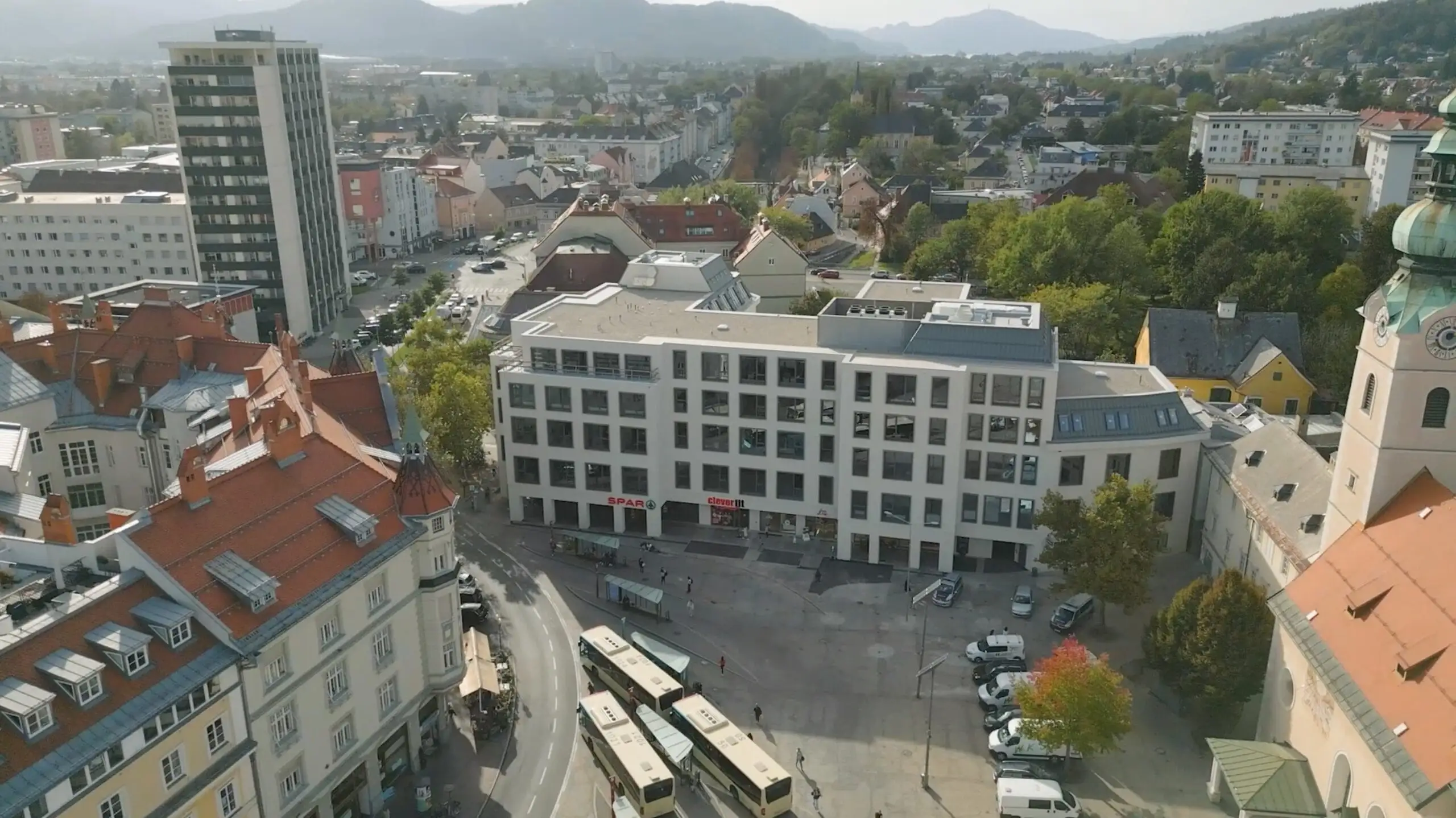 Aerial view of Klagenfurt: a square with buses and shops, with housing and the Carinthian hills behind