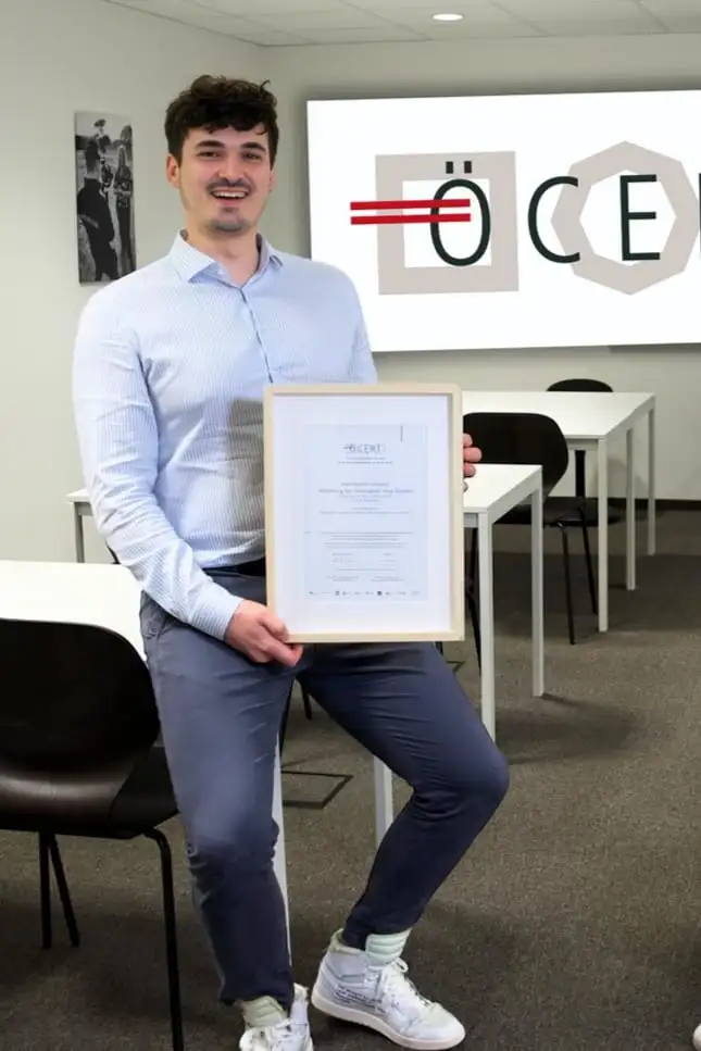 Dominik Pototschnig holding a framed ÖCERT certificate, the ÖCERT logo on a screen behind him